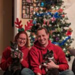 A family Christmas photo in front of a Christmas tree. Nikki and her husband Gerald and holding their grey cats who are each wearing sparkly ties.